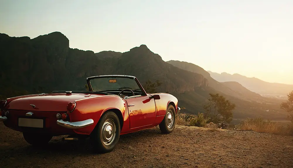 Röd cabriolet parkerad på en utsiktsplats med berg och dalgång i kvällssol.